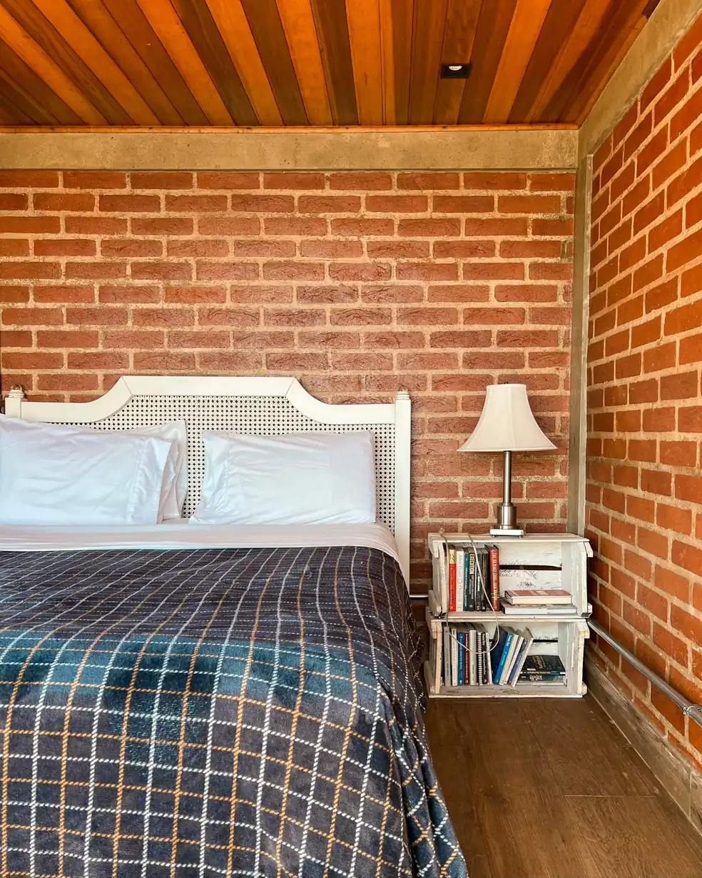 Quarto com cabeceira de palhinha, parede de tijolo aparente, teto de madeira e janela para as árvores